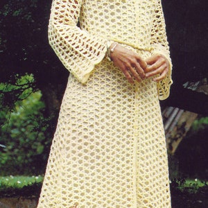 May include: A light yellow, open-weave crochet dress with a hood. The dress has long sleeves and a V-neck. The dress is floor-length and appears to be handmade. The background is a garden setting with green foliage.