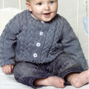 May include: A gray knitted baby cardigan with a cable knit pattern and white buttons. The cardigan is worn by a baby sitting on a white surface.