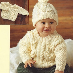 May include: Cream-coloured knitted baby sweater and hat set. The sweater features a cable knit design with textured details. The hat has a matching cable knit pattern and a small pom-pom on top. The set is displayed on a wooden surface.
