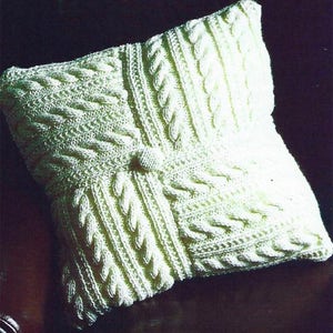 Könnte beinhalten: Ein weißes gestricktes Kissen mit einem Zopfmuster. Das Kissen ist quadratisch und hat einen Knopf in der Mitte.