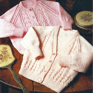 Puede incluir: Dos cárdigans de punto para bebé, uno rosa y otro crema, con un patrón de trenza y botones.