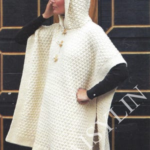May include: A white knitted hooded poncho with a textured pattern and wooden buttons. The poncho has a slit on each side and is worn over a black long-sleeved shirt.