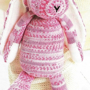 May include: A pink and white knitted bunny rabbit toy with long ears. The bunny is sitting on a cream-colored knitted surface.