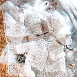 Könnte beinhalten: Ein Set weißer gestrickter Babykleidung, bestehend aus einem Cardigan, einer Mütze und Söckchen. Die Kleidung liegt auf einem Hintergrund aus Blütenblättern.