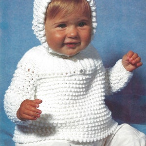 Op de afbeelding: Een witte gehaakte babytrui met bijpassende capuchon. De trui heeft een knoopsluiting en een textuurpatroon.