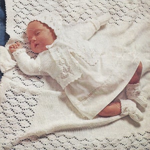 May include: A white crocheted baby blanket, dress, cardigan, and booties set. The baby is sleeping on the blanket.