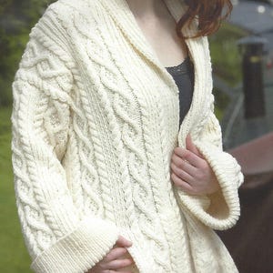 May include: A white cable knit cardigan with a shawl collar. The cardigan is open and the woman is wearing a black vest top underneath.