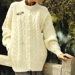 May include: Cream-colored cable-knit cardigan with a high collar and button-down front. The sweater features intricate cable patterns and is adorned with a decorative brooch. The sleeves are long, and the cardigan appears to be made of a thick, warm material.