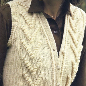 May include: Cream-colored knitted vest with a textured chevron design. The sleeveless garment features a V-neck, button closure, and a ribbed hem. The vest is worn over a brown collared shirt.