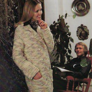 Könnte beinhalten: Eine Frau trägt einen langen, doppelreihigen Strickcardigan mit einem strukturierten Muster in Beige-, Braun- und Weißtönen. Der Cardigan hat große Knöpfe und Taschen. Die Frau steht vor einer Pflanze mit Blättern und einem Mann, der auf einem Schaukelstuhl sitzt.