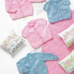 Puede incluir: Cuatro cárdigans de punto para bebé en rosa y azul con cierres de botón. Los cárdigans se muestran sobre un fondo blanco con almohadas decorativas.