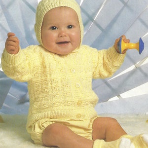 Puede incluir: Un cárdigan de punto amarillo para bebé con un gorro a juego. El cárdigan tiene cierre de botones y un patrón texturizado. El bebé lleva un pañal amarillo y calcetines blancos.