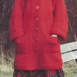 May include: A red knitted cardigan with a button fastening and two pockets. The cardigan is worn over a white turtleneck and a plaid skirt. The person is wearing brown knee-high boots.