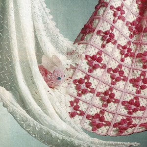 Può includere: Una copertina per neonati in crochet bianco con un bordo in crochet rosa e un motivo a fiori in crochet rosa. Un coniglietto in crochet rosa è annidato nella copertina.