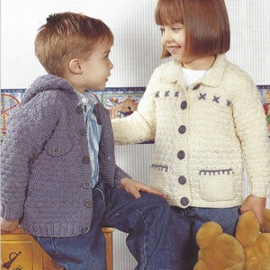 May include: Two children wearing knitted jackets. One jacket is a muted purple with a hood and large buttons. The other is cream-colored with dark buttons and decorative stitching. Both children are wearing blue jeans.