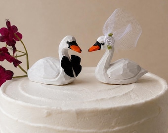 May include: Two carved white swan figurines placed on top of a frosted white cake. One swan wears a black bow tie, and the other swan wears a small white fabric veil with a flower accent. A sprig of dark purple flowers is positioned to the left.