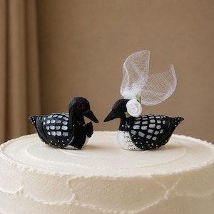 May include: Two carved wooden loon figurines placed on top of a frosted cake. One loon wears a black bow tie, while the other features a white fabric flower and a sheer white tulle veil attached to its head. Both birds have black and white plumage.
