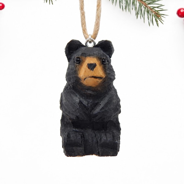 May include: A hand-carved wooden bear ornament featuring a black-painted body with a natural light brown face and snout. The bear has small dark eyes and is suspended by a tan jute twine loop attached to a silver-toned metal eyelet at the top.