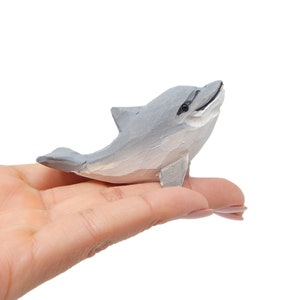 Può includere: Una piccola statuetta di delfino in legno grigio e bianco. Il delfino è in una posa di salto con la bocca aperta.