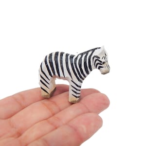 May include: A small, hand-carved wooden zebra figurine. The zebra has black and white stripes and is standing. The figurine is made of light-colored wood and is held in a hand.