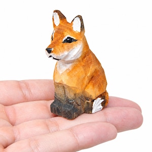 Könnte beinhalten: Eine kleine, handgeschnitzte Holzfigur eines Fuchses mit strukturiertem orange-braunem Fell und weißen Akzenten an den Ohren und der Schwanzspitze. Der Fuchs hat schwarz bemalte Augen und eine schwarze Nase sowie sichtbare Schnitzspuren.