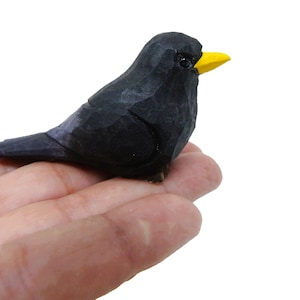 May include: A small, hand-carved wooden blackbird with a yellow beak. The bird is perched on a hand.