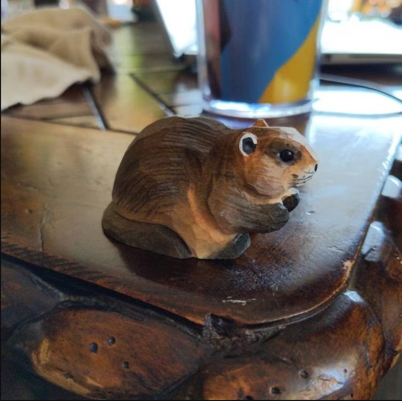 A hand-carved wooden figurine of a brown beaver sitting on a wooden surface. The beaver has a detailed face with black eyes and a small, pointed nose.