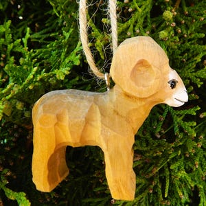 May include: A wooden carved ornament of a brown bighorn sheep with a white face. The ornament is hanging from a string.