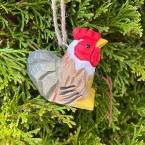 May include: A hand-carved wooden rooster ornament. It has a red comb and wattle, a yellow beak, and a gray and brown body. The rooster is on a yellow base and suspended by a natural twine string. The background is a dense, green, leafy plant.