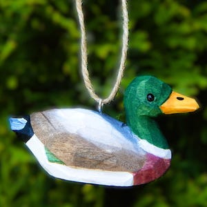 A hand-carved wooden mallard duck ornament with a green head, orange beak, and brown, white, and green body. The duck is hanging from a twine string.