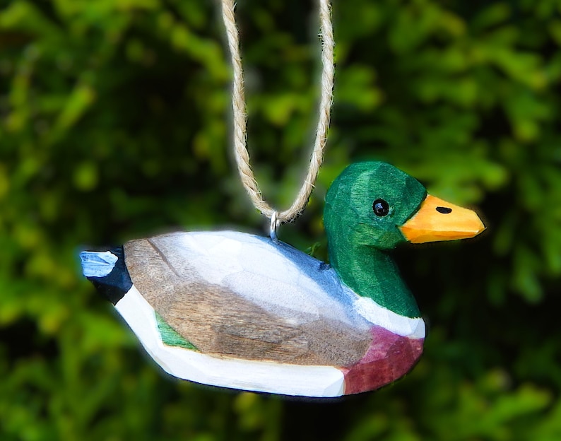 A hand-carved wooden mallard duck ornament with a green head, orange beak, and brown, white, and green body. The duck is hanging from a twine string.