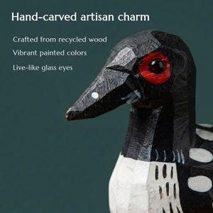 May include: A hand-carved wooden bird figurine featuring a black head and body with white spotted markings, a red patch around the dark glass eye, and a pointed beak. The text reads: Hand-carved artisan charm, Crafted from recycled wood, Vibrant painted colors, Live-like glass eyes.