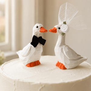 May include: Two white carved duck figurines with orange beaks and feet. One duck wears a black fabric bow tie, and the other duck wears a white fabric flower with a sheer white mesh veil attached to its head, both positioned on a white cake.