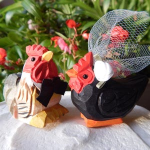 May include: A pair of wooden rooster figurines, one in black and one in white and red, with a black bow tie and a white veil. The figurines are on a white cake.