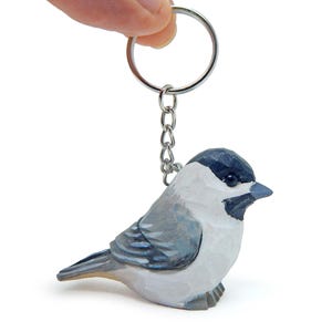May include: A hand-carved wooden keychain featuring a black-capped chickadee. The bird is painted in shades of gray, white, and black. The keychain is attached to a silver metal ring and chain.