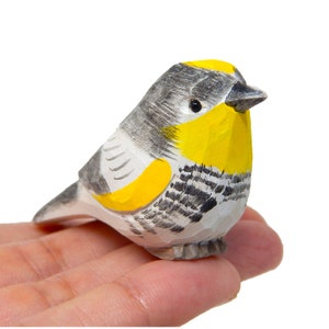 Könnte beinhalten: Eine handgeschnitzte Holzvogelfigur mit gelber Brust, grauen und weißen Flügeln und schwarzen Markierungen. Der Vogel sitzt auf einem Finger.