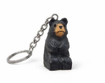 May include: A keychain featuring a small, hand-carved wooden bear figurine. The bear is painted black with a tan-colored snout and face. It has a textured, chiseled surface and is attached to a silver-toned metal chain and a circular split ring.