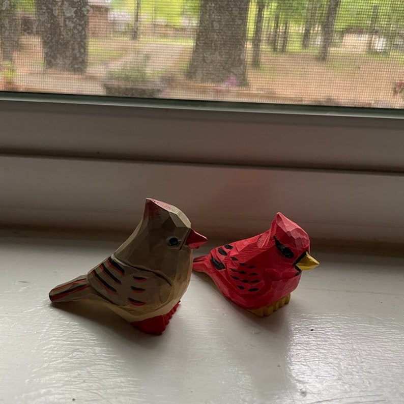 Two wooden bird figurines, one red and one brown, with painted details. The red bird has a yellow beak and black eyes. The brown bird has a black beak and black eyes. Both birds are facing the right side of the image.