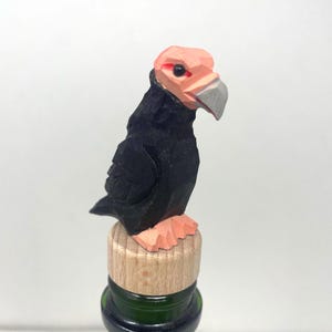 Könnte beinhalten: Ein handgeschnitzter Weinflaschenverschluss aus Holz mit einem Vogel mit schwarzem Körper, pfirsichfarbenem Kopf und Füßen sowie einem silbernen Schnabel. Der Vogel sitzt auf einem Holzverschluss, der in eine grüne Glasflasche eingesetzt ist.