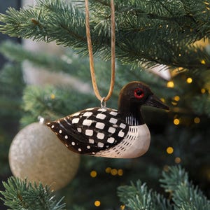 K&ouml;nnte beinhalten: Ein geschnitztes Eistaucher-Ornament aus Holz mit schwarzem Kopf, rotem Auge, schwarzem Schnabel und einem K&ouml;rper mit wei&szlig;en Quadraten und Punkten. Der Vogel h&auml;ngt an einer beigen Kordelschlaufe und zeigt detaillierte Holzmaserungen.