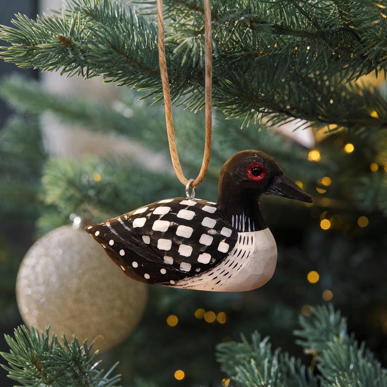 K&ouml;nnte beinhalten: Ein geschnitztes Eistaucher-Ornament aus Holz mit schwarzem Kopf, rotem Auge, schwarzem Schnabel und einem K&ouml;rper mit wei&szlig;en Quadraten und Punkten. Der Vogel h&auml;ngt an einer beigen Kordelschlaufe und zeigt detaillierte Holzmaserungen.
