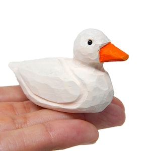 Könnte beinhalten: Eine weiße Holz-Entenfigur mit orangefarbenem Schnabel. Die Ente ist mit einer detaillierten Textur geschnitzt und sitzt auf einer Hand.
