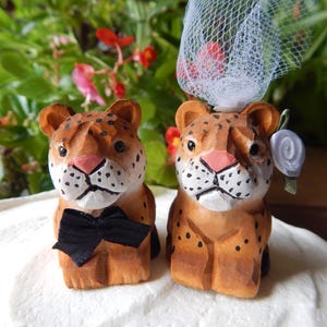 May include: A pair of wooden leopard figurines, one wearing a black bow tie and the other a white veil and a blue flower. The figurines are brown with black spots and are standing on a white surface.
