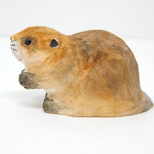 A hand-carved wooden beaver figurine with a light brown finish. The beaver is sitting on its haunches with its tail behind it.