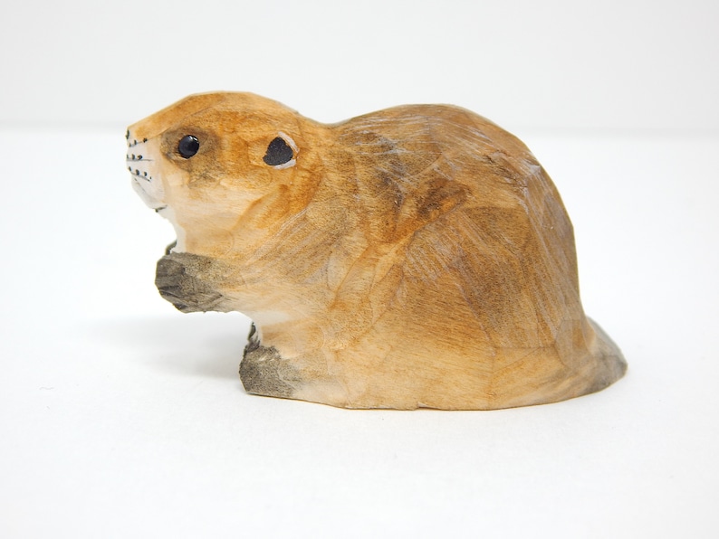 A hand-carved wooden beaver figurine with a light brown finish. The beaver is sitting on its haunches with its tail behind it.
