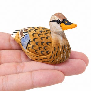 Könnte beinhalten: Eine kleine, handgeschnitzte Entenfigur aus Holz mit beigem Körper und schwarzen Federzeichnungen, einem leuchtend orangefarbenen Schnabel mit schwarzen Akzenten und einem blauen Fleck am Flügel. Die Ente hat eine glatte Struktur und ein kleines schwarzes Auge.