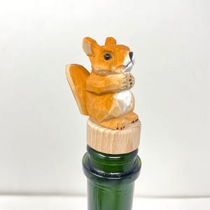 Puede incluir: Un tapón de botella de madera con una ardilla tallada con pelaje naranja y blanco. La ardilla está posada sobre un corcho, que está colocado en una botella de vidrio verde. La ardilla está juntando sus patas.