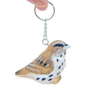 May include: A wooden bird keychain with brown and white painted details. The bird is perched on a silver key ring.