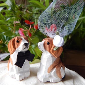 Pode incluir: Um topo de bolo de casamento de madeira com dois cães beagle. O cão noivo está usando uma gravata borboleta preta e a cadela noiva está usando um véu branco. Os cães estão em pé sobre uma base de bolo branca.