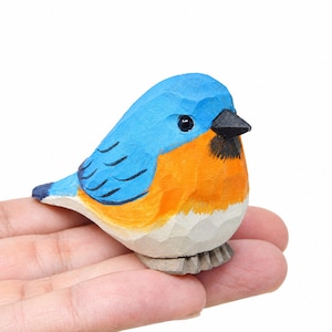 May include: A small hand-carved wooden bird figurine featuring a bright blue head and back, an orange chest, and a white belly. The bird has a black beak, a dark eye, and visible carved facets throughout its surface, showcasing a textured finish.